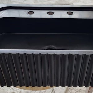 May include: A black rectangular sink with a fluted, ridged apron. The sink has a rectangular basin and a raised area with three tap holes. The sink is made of a dark material and is likely for a bathroom or kitchen.