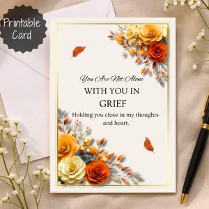 May include: A sympathy card with the text "You Are Not Alone WITH YOU IN GRIEF". The card features floral arrangements with orange, yellow, and white roses. A black pen with gold accents rests beside the card.