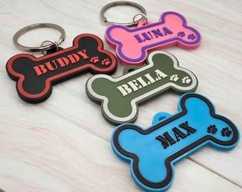 May include: Four bone-shaped keychains with metal rings. One is black with red text BUDDY, one pink with purple text LUNA, one olive green with white text BELLA, and one blue with black text MAX. Each features small paw print details on the right side.