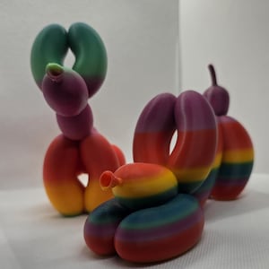 Puede incluir: Tres esculturas de animales de globos de colores. Las esculturas tienen la forma de un perro, un caracol y un animal de globo. Los colores incluyen rojo, naranja, amarillo, verde y morado. Las esculturas están impresas en 3D.