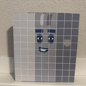 May include: A square box with a grid pattern in shades of gray, featuring a cartoon face with blue eyes, eyebrows, and a smiling mouth. The box is sitting on a white surface.