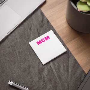 May include: A stack of white sticky notes with the letters "MCM" in bright pink. The notes are on a dark gray surface, with a laptop and pen visible in the background. The scene is set on a wooden desk.