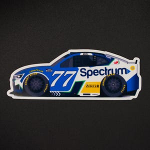 May include: A sticker depicting a race car with the number 77 in blue and white. The car has sponsor logos, including "Spectrum" and "Goodyear." The car is mainly blue, white, and green, with black tyres.