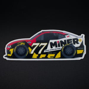 May include: A sticker depicting a race car in black, red, and white. The number "77" is in yellow, and the word "MINER" is displayed prominently. The design includes sponsor logos and the text "DOCKS, DOORS AND MORE."