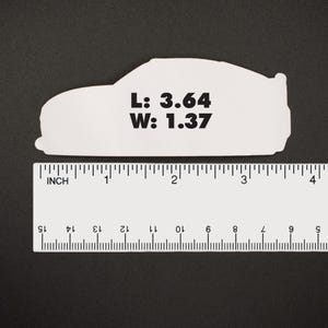 May include: A white car-shaped sticker with the text "L: 3.64 W: 1.37" printed on it. The sticker is measured with a ruler in inches. The car shape is a side profile of a coupe.