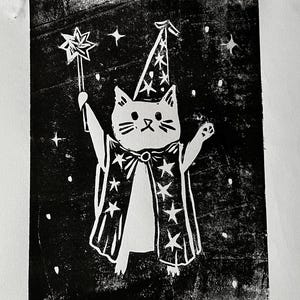 May include: A black and white print of a cat dressed as a wizard, holding a star-tipped wand. The cat wears a pointed hat and a cape adorned with stars. The background is black with white stars.