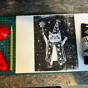 May include: A linocut print of a cat wizard holding a wand, with stars, on white paper. Includes a red cutting tool, a red cone, a pencil, and a tube of black Essdee Lino Ink for block printing.