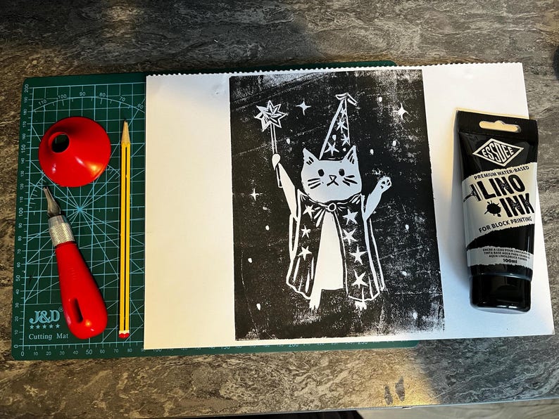 May include: A linocut print of a cat wizard holding a wand, with stars, on white paper. Includes a red cutting tool, a red cone, a pencil, and a tube of black Essdee Lino Ink for block printing.