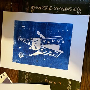 May include: A white linocut print on paper featuring a cat dressed as a wizard against a blue background. The cat wears a wizard hat and cape adorned with stars, holding a star-tipped wand. The print includes stars scattered around the cat.