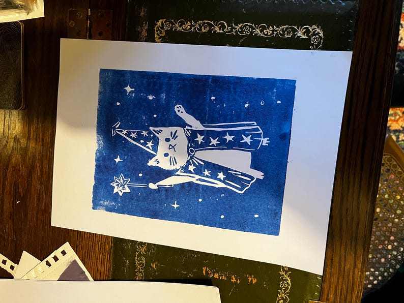 May include: A white linocut print on paper featuring a cat dressed as a wizard against a blue background. The cat wears a wizard hat and cape adorned with stars, holding a star-tipped wand. The print includes stars scattered around the cat.