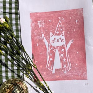 May include: A white cat illustration in a wizard costume, holding a star wand, printed on pink paper. The cat wears a pointed hat and a cape with star details. The print is on a white background.