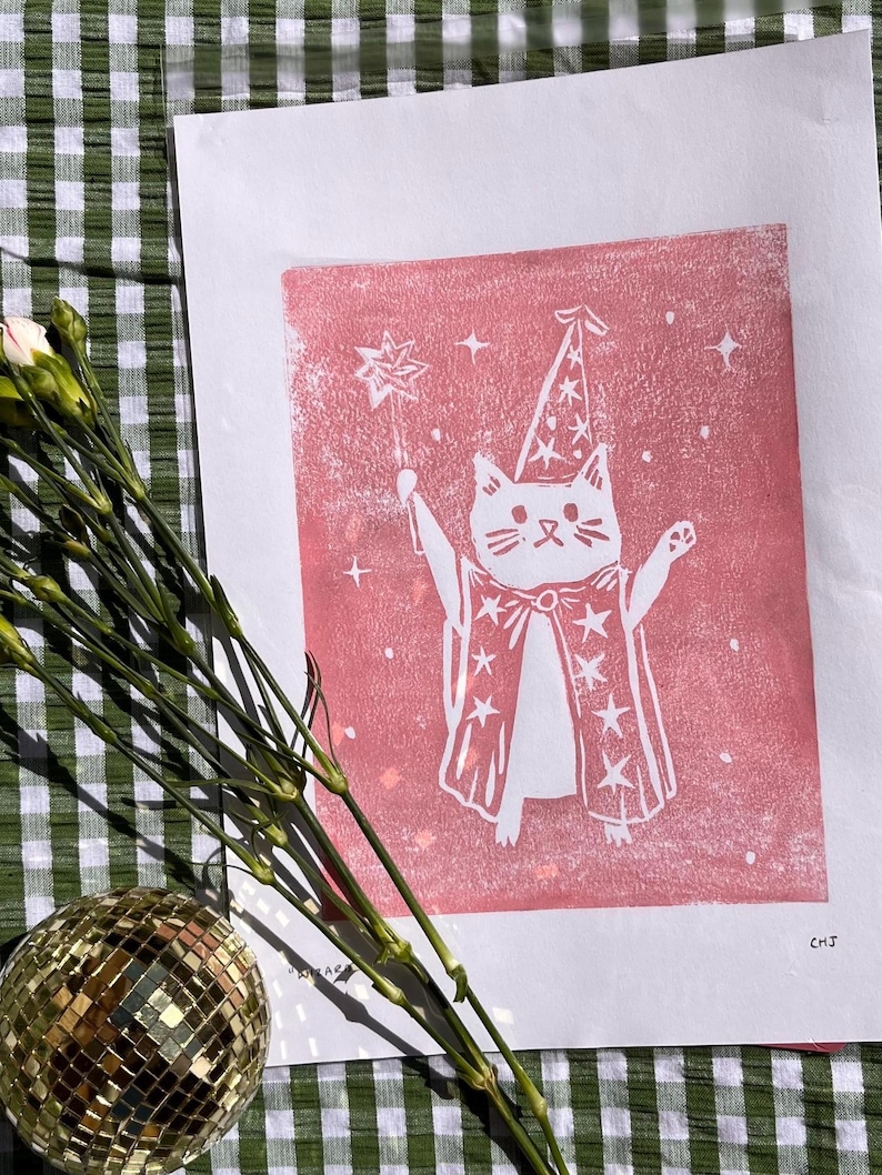 May include: A white cat illustration in a wizard costume, holding a star wand, printed on pink paper. The cat wears a pointed hat and a cape with star details. The print is on a white background.