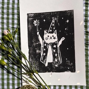 May include: A black and white print of a cat dressed as a wizard, holding a star-topped wand. The cat wears a wizard hat and cape with star designs. The print is on white paper, with a green and white checkered background.