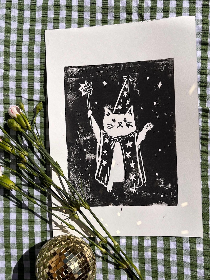 May include: A black and white print of a cat dressed as a wizard, holding a star-topped wand. The cat wears a wizard hat and cape with star designs. The print is on white paper, with a green and white checkered background.