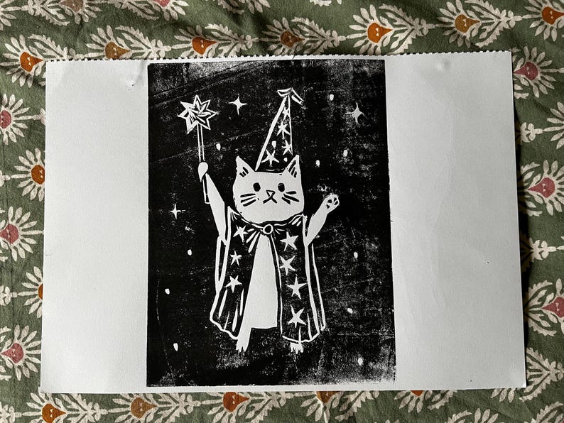 May include: A black and white print of a cat dressed as a wizard, holding a star-tipped wand. The cat wears a pointed hat and a cape adorned with stars. The background is black with white stars.