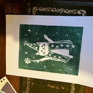 May include: A white cat wearing a wizard hat and robe, holding a star-tipped wand, printed on a dark green background with white stars. The cat illustration is on a white rectangular paper.