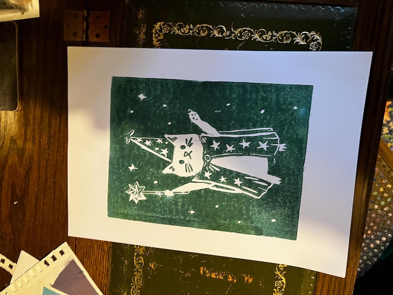 May include: A white cat wearing a wizard hat and robe, holding a star-tipped wand, printed on a dark green background with white stars. The cat illustration is on a white rectangular paper.