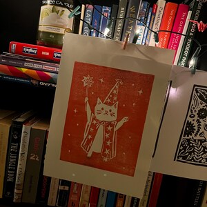 May include: A red and white print of a cat wizard with a star-topped wand and cape, hanging on a string with a wooden clip. The print is displayed in front of a bookshelf filled with books, a candle, and string lights.