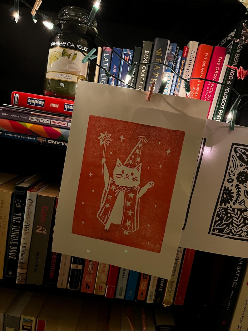 May include: A red and white print of a cat wizard with a star-topped wand and cape, hanging on a string with a wooden clip. The print is displayed in front of a bookshelf filled with books, a candle, and string lights.