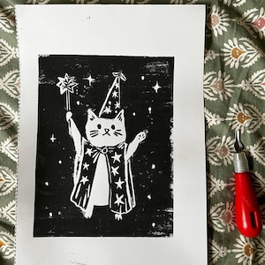 May include: A black and white linocut print depicting a cat wizard with a star wand. The cat wears a wizard hat and cape adorned with stars. A red carving tool is visible in the image.