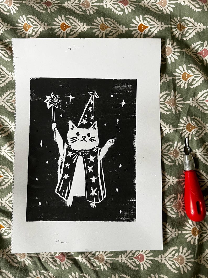 May include: A black and white linocut print of a cat dressed as a wizard, holding a star wand. The cat wears a wizard hat and cape with star details. A red carving tool is visible on the right.