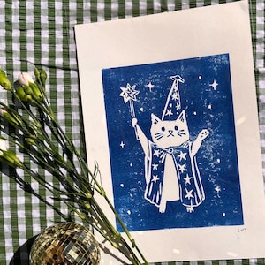 May include: A white cat in a wizard costume with a star wand, printed on blue paper. The cat is wearing a pointed hat and a cape with star designs. The print is on a white background, with a green and white checkered cloth in the background.