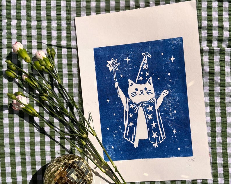 May include: A white cat in a wizard costume with a star wand, printed on blue paper. The cat is wearing a pointed hat and a cape with star designs. The print is on a white background, with a green and white checkered cloth in the background.