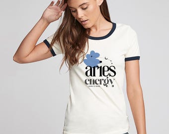 Puede incluir: Camiseta ringer color crema con ribete azul marino. La camiseta presenta las palabras "aries energy" en negro, con un diseño floral azul y el texto "BORN TO BLAZE". La modelo lleva pantalones color turquesa.