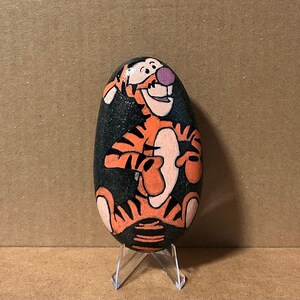 May include: A hand-painted, oval-shaped rock featuring the character Tigger from Winnie the Pooh. Tigger is depicted in orange and black, with a pink nose and a smiling expression. The rock is set against a neutral background.