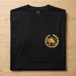 May include: Black t-shirt with a gold emblem of a lion within a wreath. The emblem is positioned on the left chest. The shirt is neatly folded on a light wood surface.