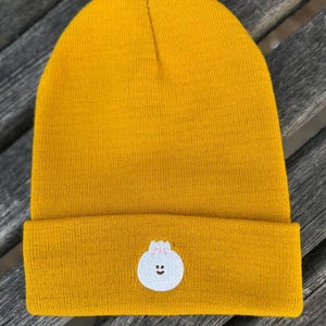 May include: A mustard yellow knit beanie featuring a cuffed brim and a small, embroidered white dumpling character on the front. The dumpling has a simple brown line for a mouth, two small brown dots for eyes, and light pink vertical lines on top.