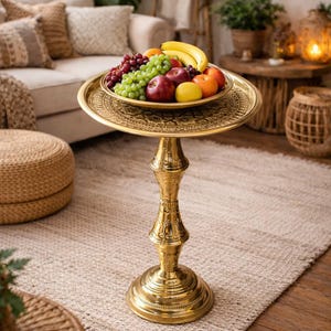 May include: A decorative gold-coloured side table with a round top, displaying a brass tray laden with fresh fruit. The fruit includes red apples, green grapes, bananas, and oranges. The table is on a neutral-toned rug.