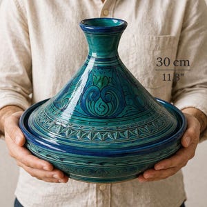 May include: A teal and blue ceramic tagine pot with a conical lid and a wide base. The pot features intricate patterns and is approximately 11.8 inches in diameter. The tagine is held by a person.