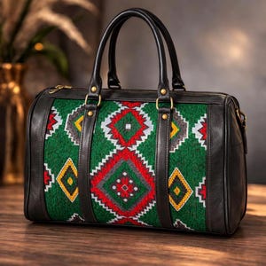 Peut inclure: Sac de voyage en cuir noir avec un panneau central orné d'un motif géométrique vert, rouge, jaune et blanc. Il possède deux poignées noires, une fermeture éclair dorée et des détails dorés. Le sac est posé sur une surface en bois.