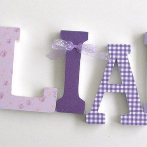 May include: Purple, pink, and white wooden letters spelling the name "LLIANA" with decorative accents, including a yellow bow, a purple bow, and a butterfly.