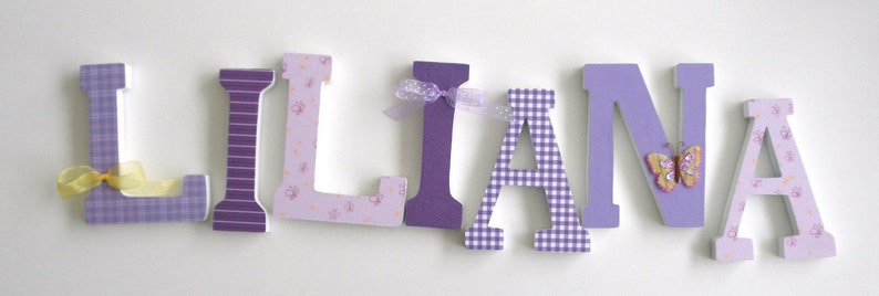 May include: Purple, pink, and white wooden letters spelling the name "LLIANA" with decorative accents, including a yellow bow, a purple bow, and a butterfly.