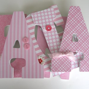 Op de afbeelding: Roze houten letters die "JADA" spellen met verschillende patronen, waaronder stippen, strepen en swirls. Elke letter heeft een roze knoop en een klein roze bloemetje.