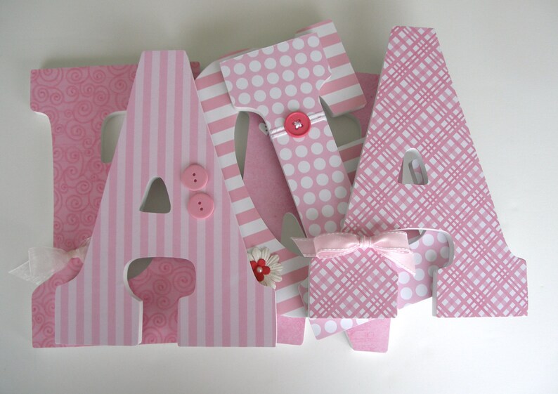 Op de afbeelding: Roze houten letters die "JADA" spellen met verschillende patronen, waaronder stippen, strepen en swirls. Elke letter heeft een roze knoop en een klein roze bloemetje.