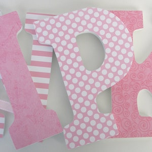 Op de afbeelding: Drie roze houten letters, A, P en K, met verschillende patronen. De A heeft roze en witte strepen, de P heeft roze met witte stippen en de K heeft een roze swirl-patroon. De letters zijn in een rij gerangschikt.