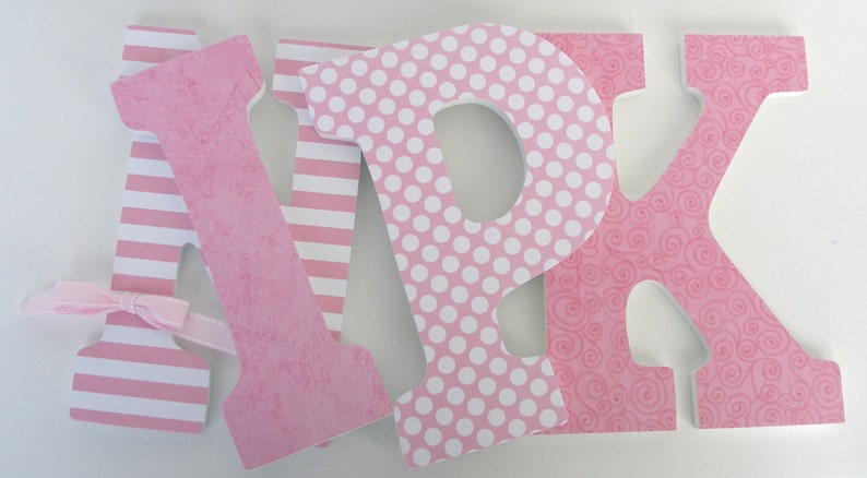 Op de afbeelding: Drie roze houten letters, A, P en K, met verschillende patronen. De A heeft roze en witte strepen, de P heeft roze met witte stippen en de K heeft een roze swirl-patroon. De letters zijn in een rij gerangschikt.