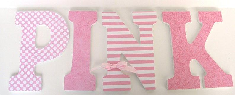 Op de afbeelding: Roze houten letters met verschillende patronen. De letter P heeft witte stippen, de letter I heeft een roze en wit textuurpatroon, de letter N heeft roze en witte strepen en de letter K heeft een roze bloemenpatroon.