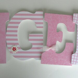 Op de afbeelding: Roze houten letters die de naam "Angela" spellen met decoratieve accenten. Elke letter heeft een ander patroon, waaronder strepen, stippen en een geruit ontwerp. De letters zijn versierd met kleine roze en witte bloemen en knopen.