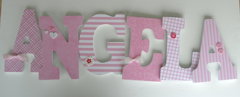 Op de afbeelding: Roze houten letters die de naam "Angela" spellen met decoratieve accenten. Elke letter heeft een ander patroon, waaronder strepen, stippen en een geruit ontwerp. De letters zijn versierd met kleine roze en witte bloemen en knopen.