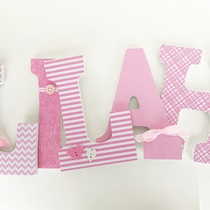 Op de afbeelding: Roze houten letters die de naam "LIAM" spellen met verschillende gepatroneerd papier dat elke letter bedekt. De letters hebben decoratieve accenten zoals knopen, strikken en harten.