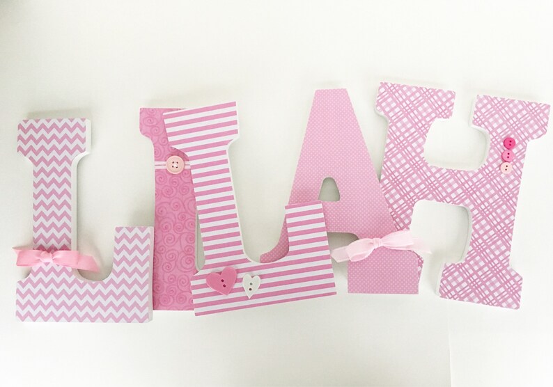 Op de afbeelding: Roze houten letters die de naam "LIAM" spellen met verschillende gepatroneerd papier dat elke letter bedekt. De letters hebben decoratieve accenten zoals knopen, strikken en harten.