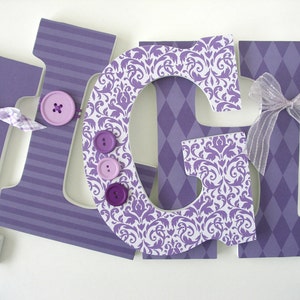 May include: Purple wooden letters spelling "GIGI" decorated with purple and white patterned paper, buttons, and a sheer purple bow.