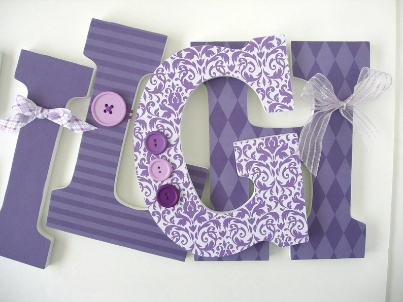 May include: Purple wooden letters spelling "GIGI" decorated with purple and white patterned paper, buttons, and a sheer purple bow.