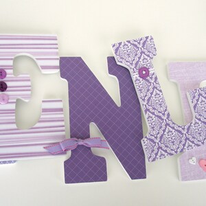 May include: Purple and white wooden letters spelling the name "KENLY" with decorative fabric and buttons. The letters are decorated with different patterns, including stripes, damask, and plaid.