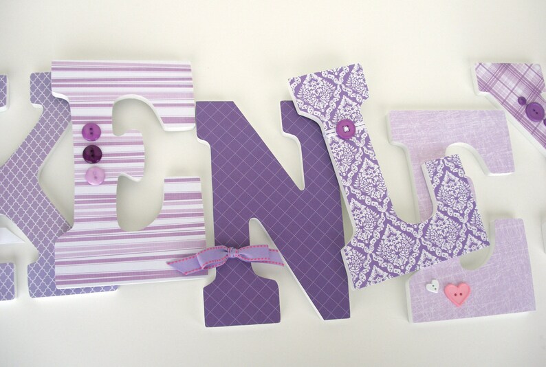 May include: Purple and white wooden letters spelling the name "KENLY" with decorative fabric and buttons. The letters are decorated with different patterns, including stripes, damask, and plaid.