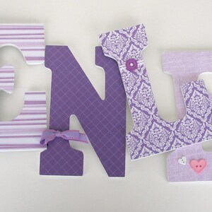 May include: Purple and white fabric letters spelling the name "KENLY" with decorative buttons and bows. The letters are made of wood and have a 3D effect.
